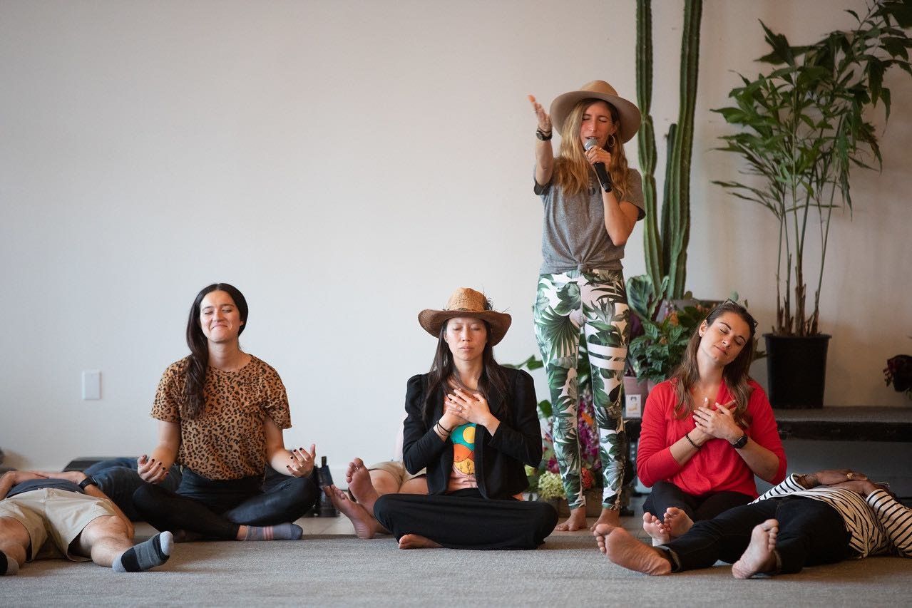Women seated and lying on the floor in a guided breathwork and sound session, one standing and singing into a microphone